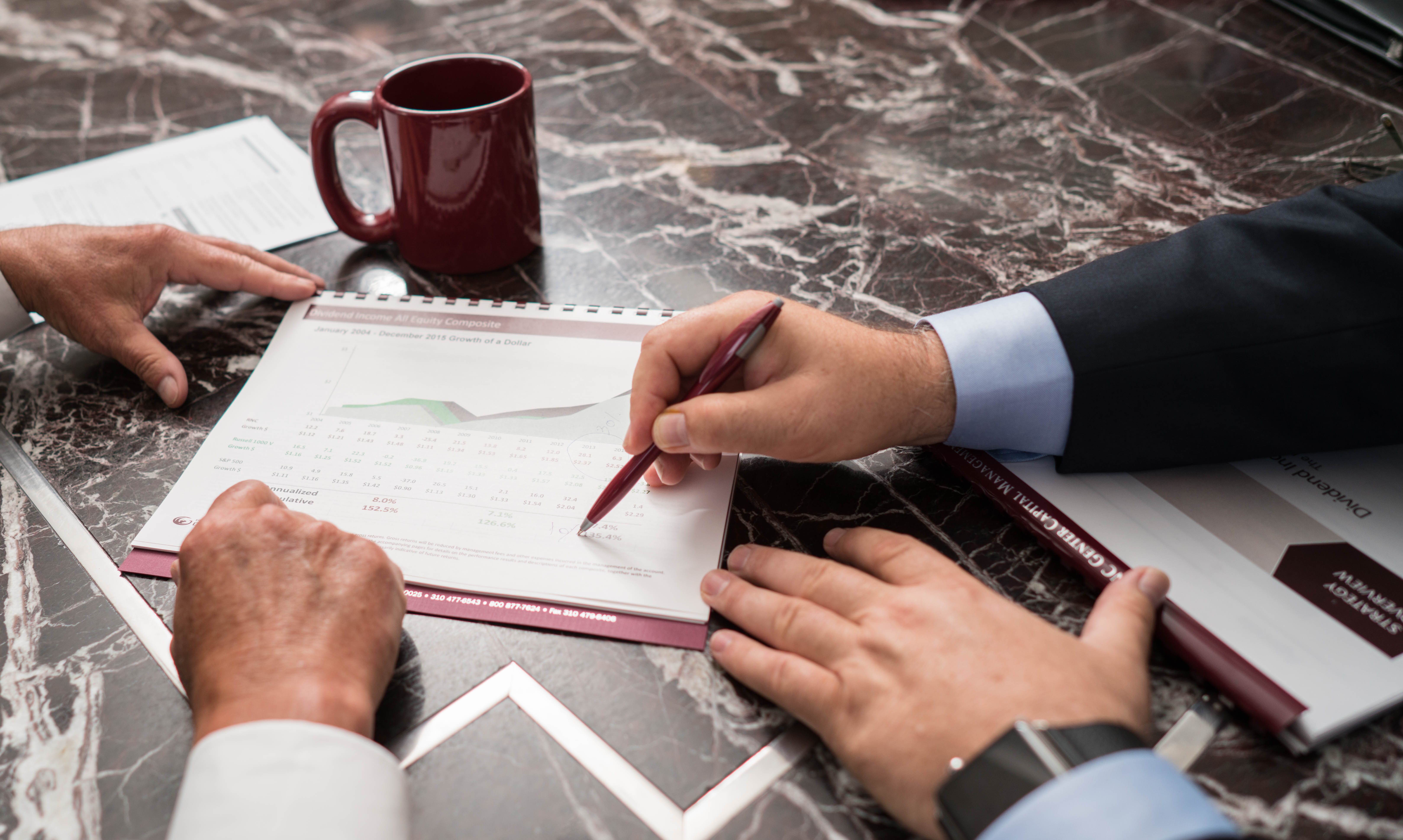 An advisor pointing with a pen at a printed performance chart while reviewing it with a client across a marble conference table